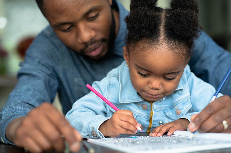 4 year old African American girl writing while father looks on