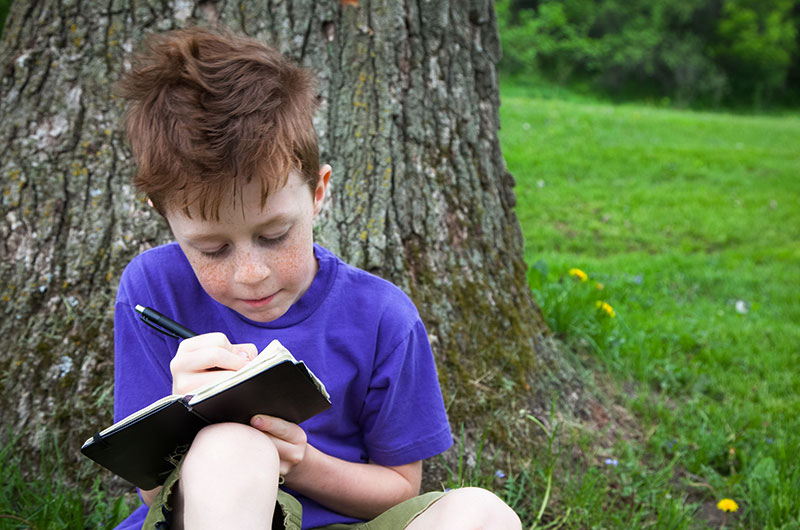 7 year old boy with red hair writing in a journal outside