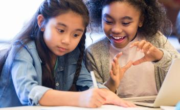 two elementary girls talking and writing in class