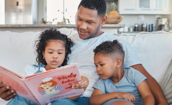 Father at home on couch reading picture book to young daughter and son
