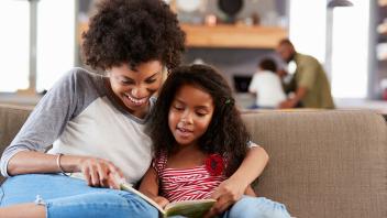 Mother reading book aloud to elementary aged daughter at home on the couch