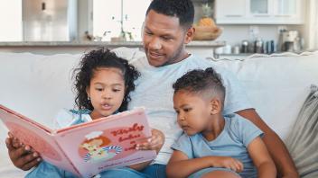 Father at home on couch reading picture book to young daughter and son