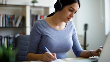 young woman wearing headphones and taking notes while working on laptop
