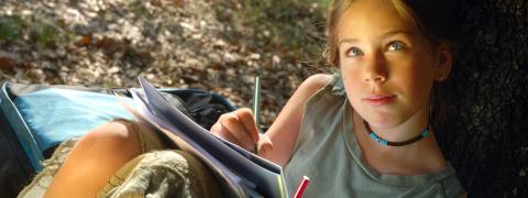 girl looks up from her writing notebook while leaning against tree outdoors